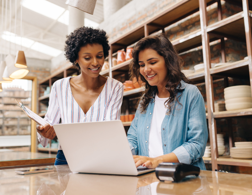 Two women smiling while logging into their online business banking acccount.  In the foreground there is a cell phone, credit card terminal and a laptop.  One of the women is holding a clipboard.  The background contains shelving with items